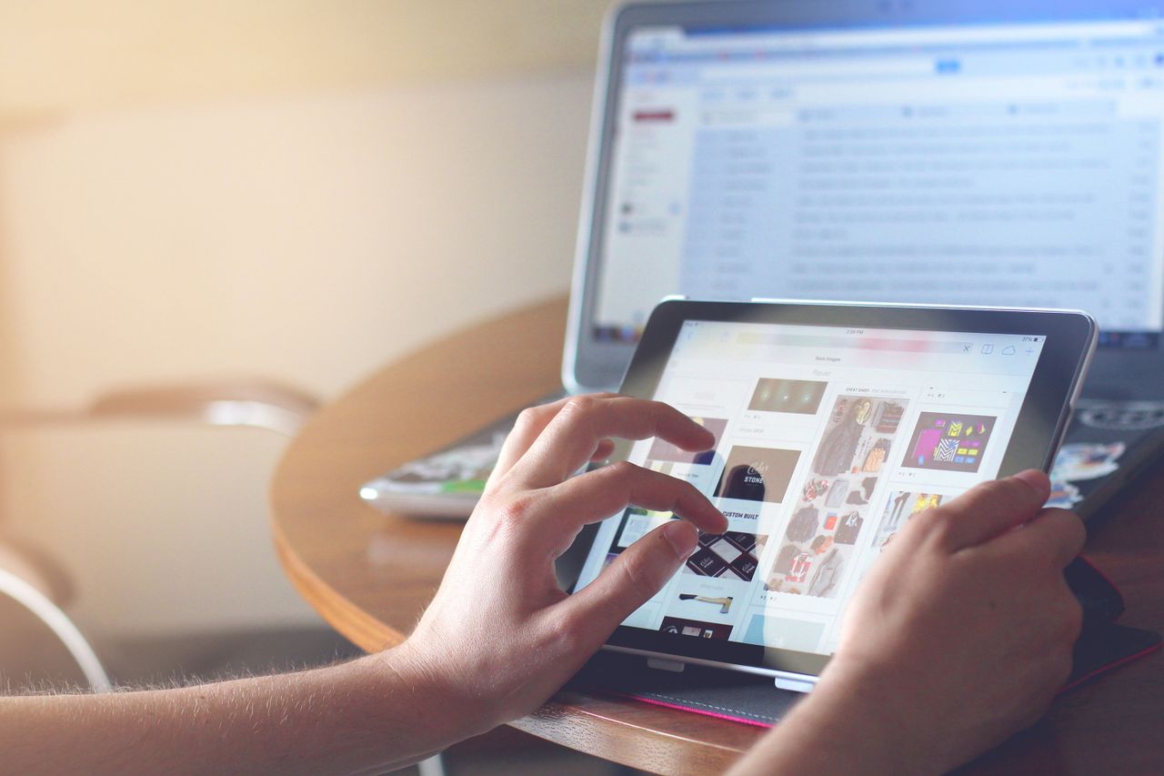 home-img Hands using a tablet beside a laptop in a home office setting, showcasing online browsing.